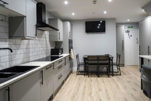A modern kitchen with grey cabinetry, a black range hood, a microwave, a dining table with chairs, and a flat-screen TV mounted on the wall.