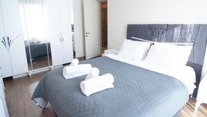 Una cama bien hecha con un edredón gris, dos toallas blancas enrolladas y un cabecero gris.