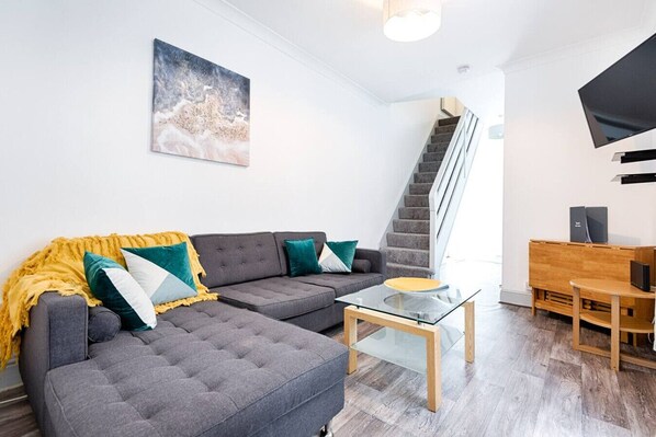 A modern living room with a grey sofa, a glass coffee table, a wooden side table, and a staircase leading to another level.