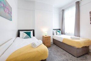 Habitación de hotel con dos camas, una mesita de noche, un ventanal con cortinas y un cuadro en la pared.