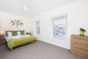 Une chambre à coucher comprenant un lit, des tables de chevet, une commode, un élément de décoration murale et des fenêtres équipées de stores.