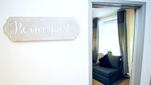 En skylt på väggen med texten ”Var vår gäst”, en mörkfärgad soffa med ett mönstrat prydnadskudd och en skymt av ett rum med ett fönster och gardiner.