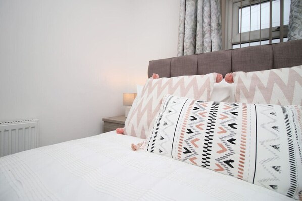 A bedroom with a bed, pillows, a nightstand, a lamp, a window with curtains, and a radiator.