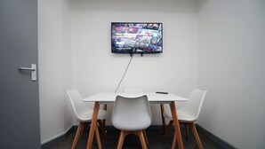 Ett litet mötesrum med ett vitt bord och fyra stolar, en platt-TV som är monterad på väggen och en dörr på vänster sida.