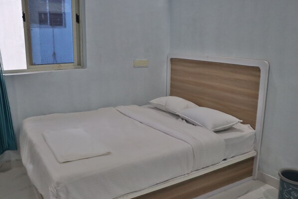 A single bed with a wooden headboard, white bedding, and a small window with a view of a building.
