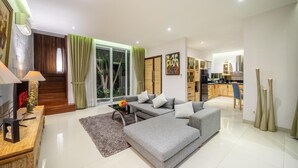 A modern living room with a grey sofa, a coffee table, and a TV on a wooden stand. There is a staircase leading to a balcony with green curtains. - Villa Nora by Five Elements (Canggu)