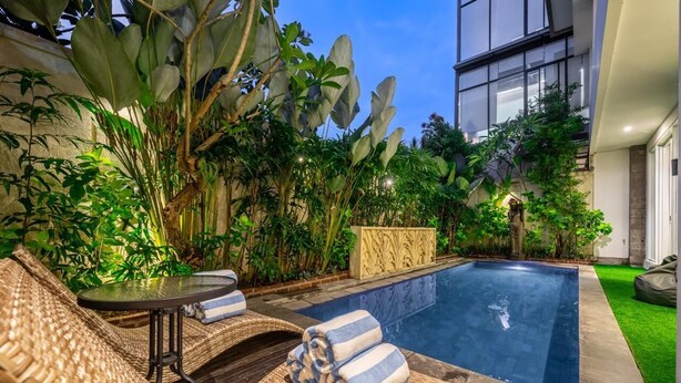 A swimming pool with lounge chairs and a table, surrounded by lush greenery and a modern building in the background. - Villa Nora by Five Elements (Canggu)
