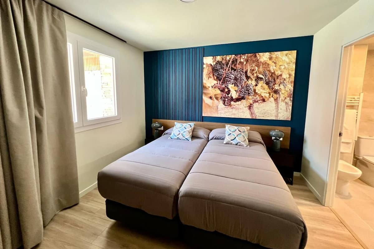 A double bed with two pillows, a window with curtains, a wall-mounted artwork of grapes, and a wooden floor.