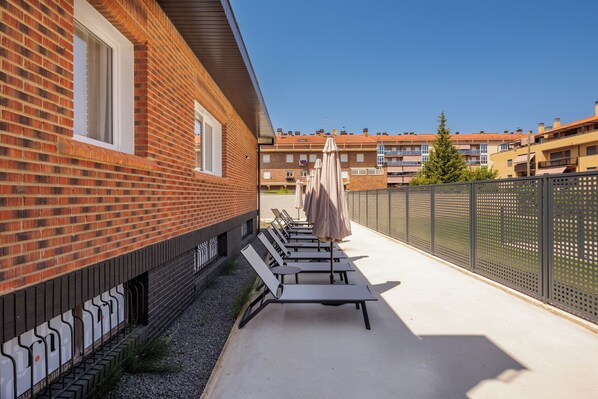 A row of sun loungers with umbrellas on a concrete patio, adjacent to a brick building with a window.