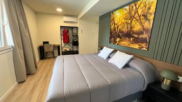 A neatly arranged hotel room with a large bed, a desk, and a chair. There is a coat rack with a red jacket, a wall-mounted air conditioner, and a large painting of autumn leaves.