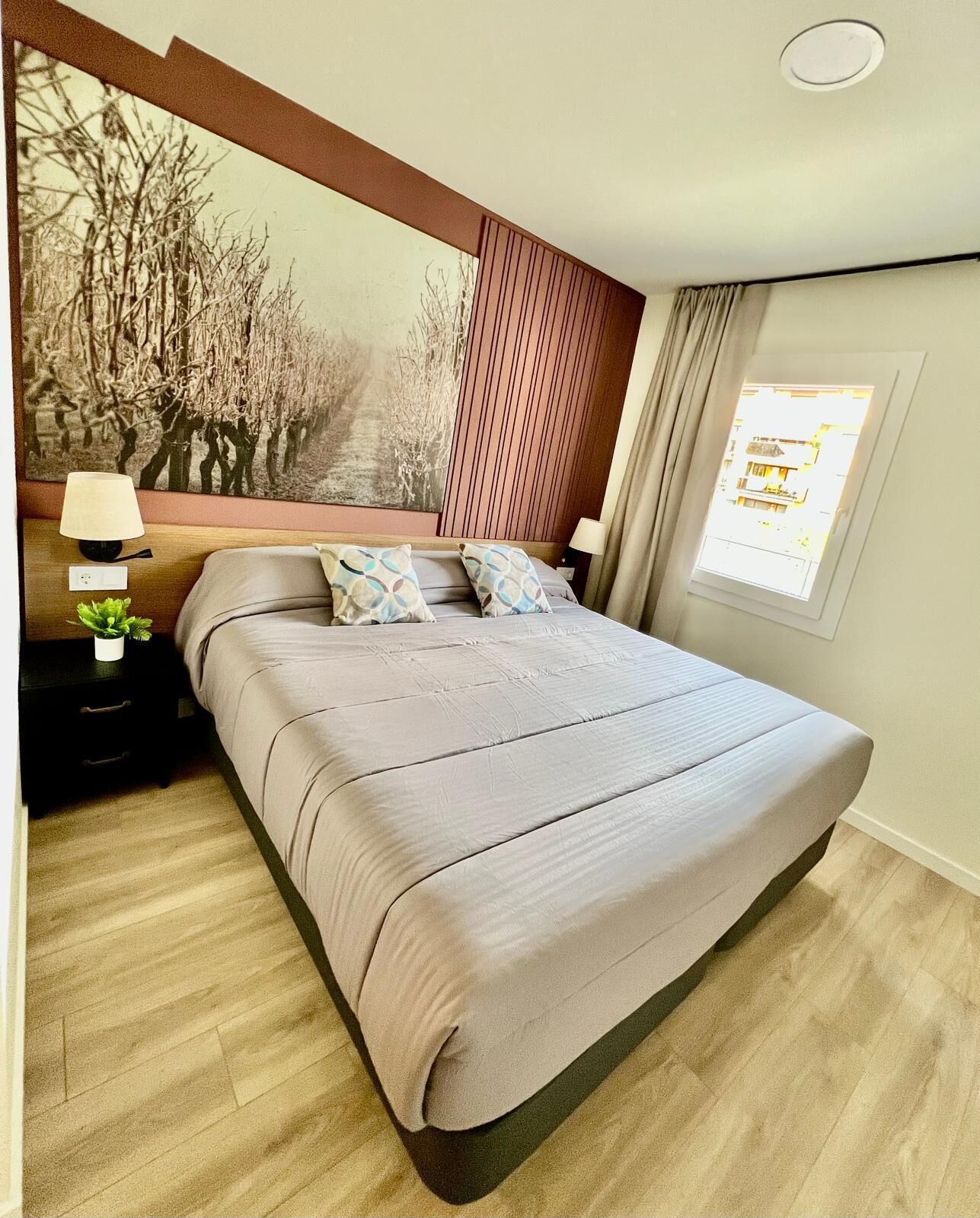 Ein modernes Schlafzimmer mit einem großen Bett, einem Nachttisch mit Lampe, einem an der Wand montierten Fernseher und einem Fenster mit Vorhängen.