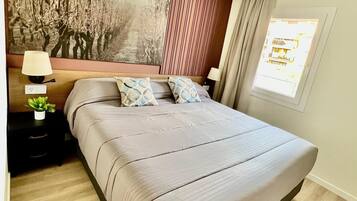 Quarto moderno com uma cama grande, uma mesa de cabeceira com abajur, TV fixada na parede e janela com cortinas.