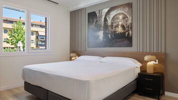 Un dormitorio moderno con una cama grande, una mesita de noche con una lámpara, una ventana con vistas a edificios y un cuadro grande en la pared.