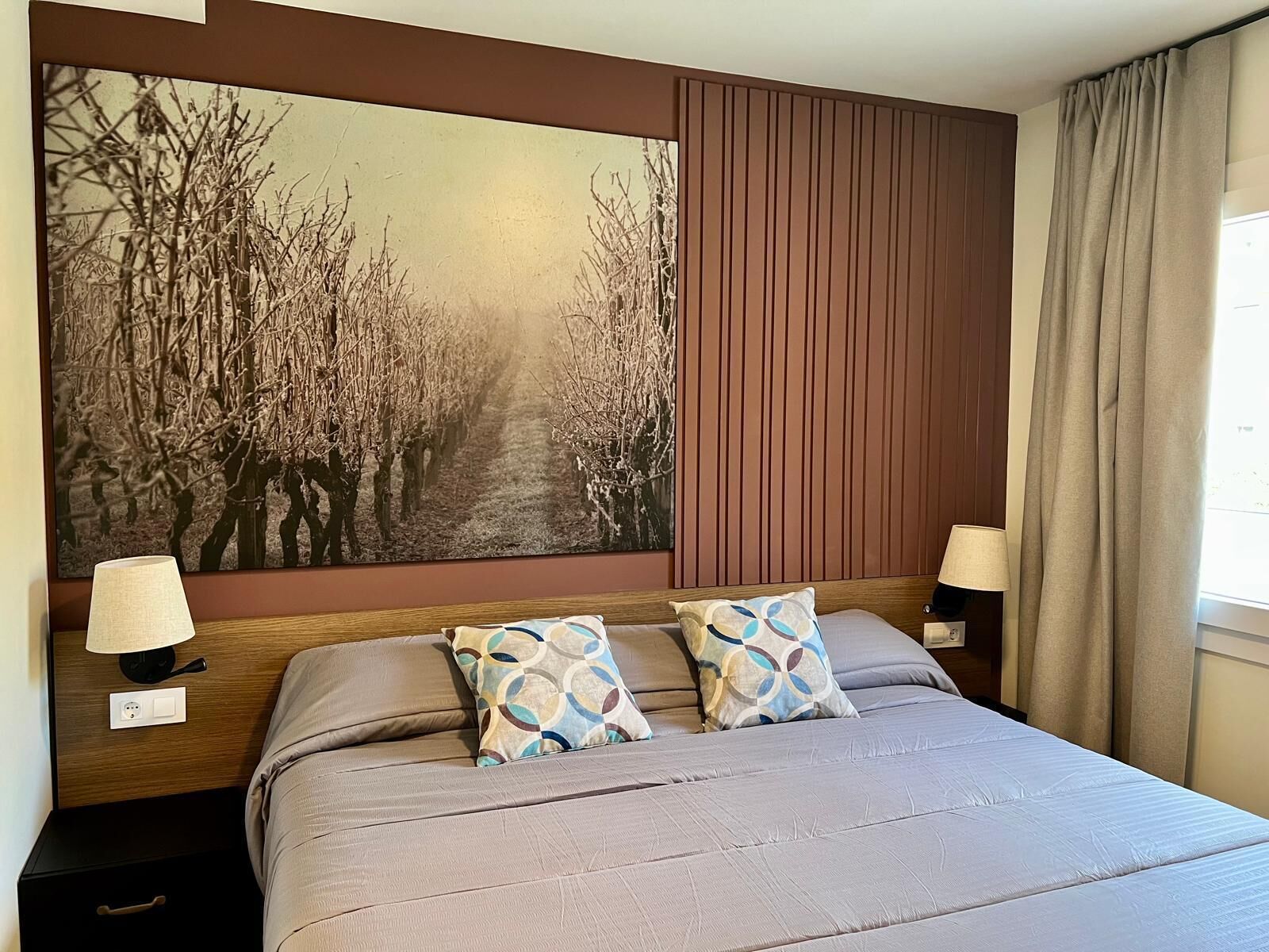 A hotel room with a large painting of a vineyard, a bed with a grey bedspread, two patterned pillows, and a nightstand with a lamp and a power outlet.