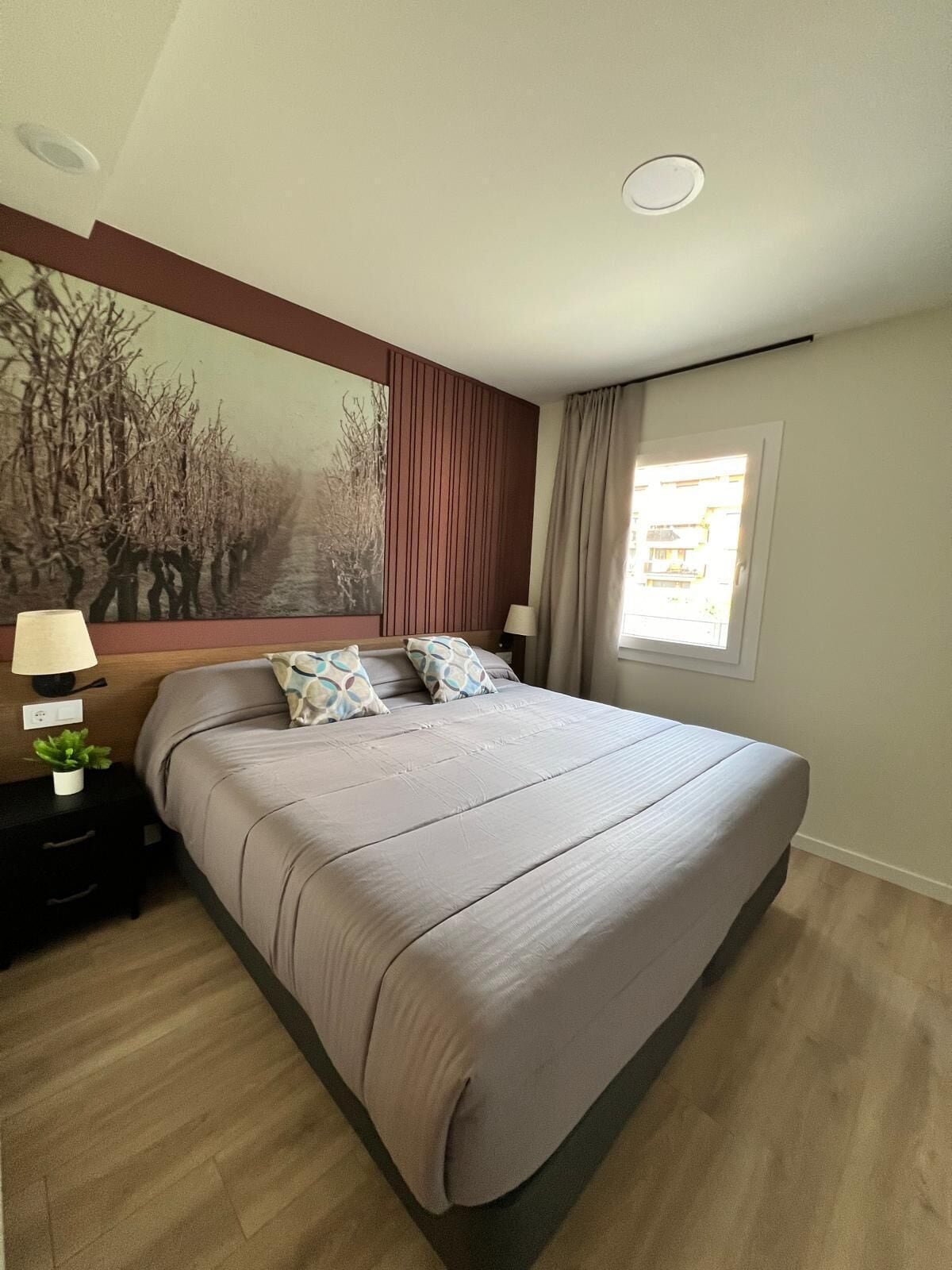 A bedroom with a large bed, a nightstand with a lamp, a wall-mounted painting, and a window with curtains.