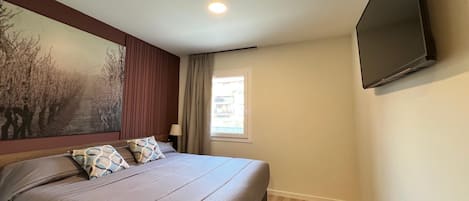 A bedroom with a bed, a TV mounted on the wall, a window with curtains, and a painting on the wall.