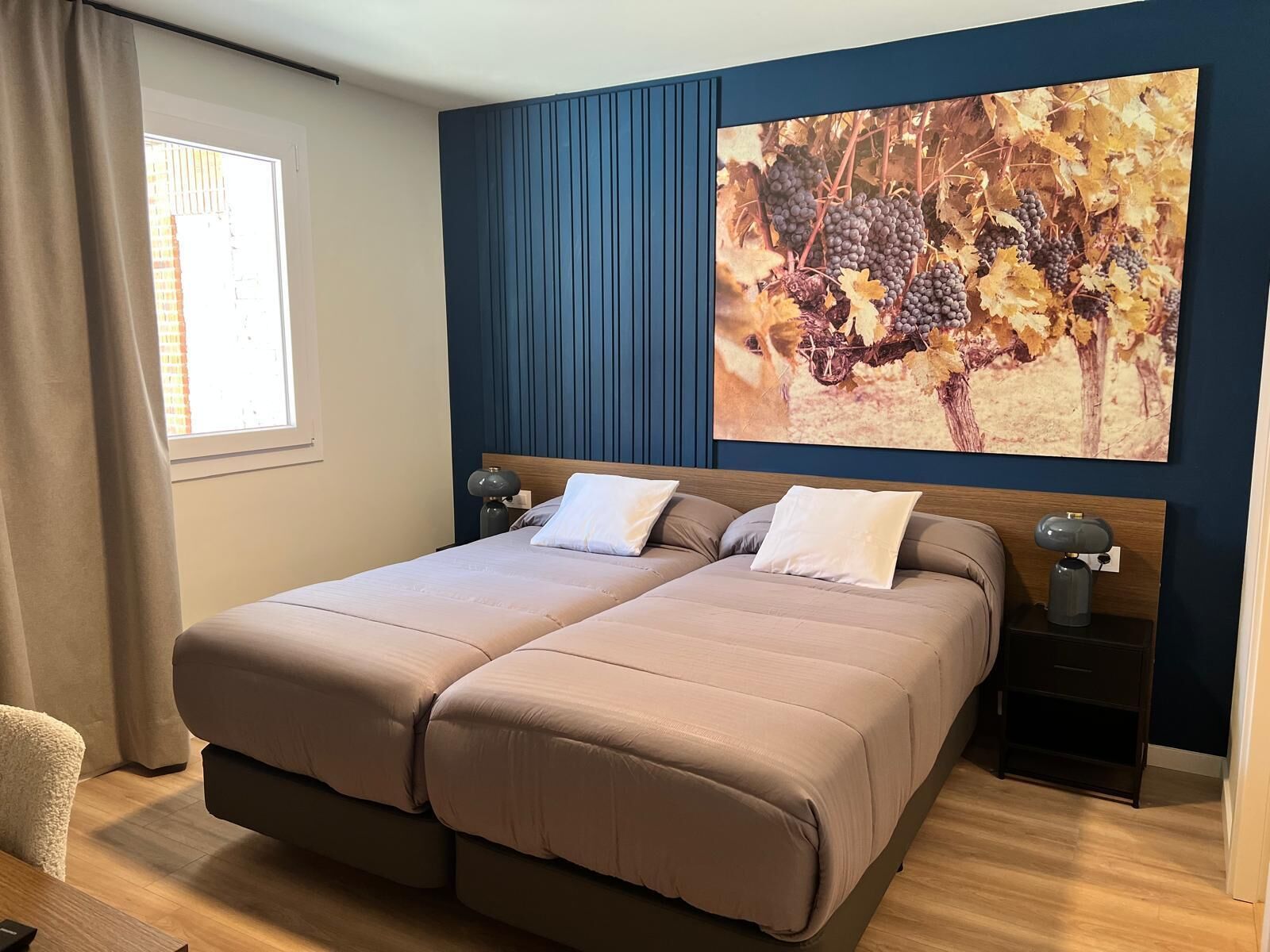 Una cama doble con cabecera, dos mesitas de noche, una ventana con cortinas y un cuadro colgado en la pared con racimos de uvas.