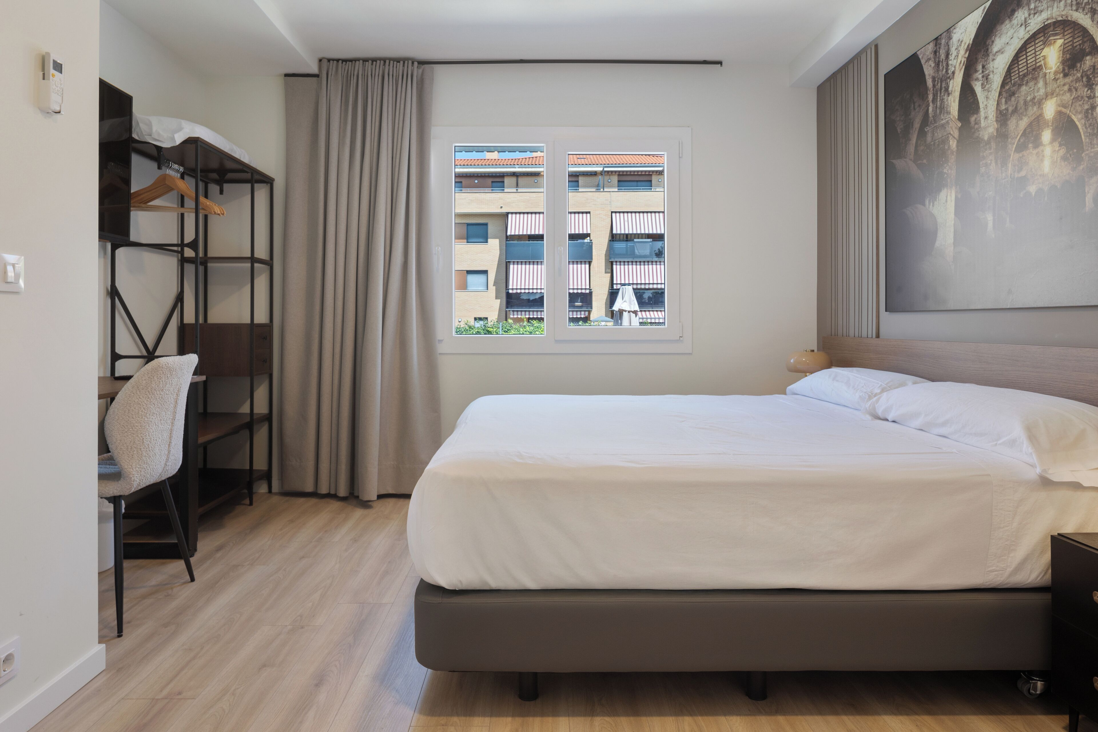 Ein Hotelzimmer mit Bett, Schreibtisch mit Stuhl, Kleiderschrank, Fenster mit Blick auf Gebäude und einem Bild an der Wand.