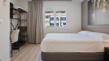 Ein Hotelzimmer mit Bett, Schreibtisch mit Stuhl, Kleiderschrank, Fenster mit Blick auf Gebäude und einem Bild an der Wand.