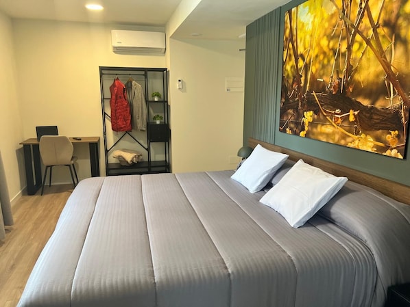 A hotel room with a bed, a desk, a chair, a coat rack, and a wall-mounted air conditioner. - Iraipe Haro Auto Check-in (Haro)