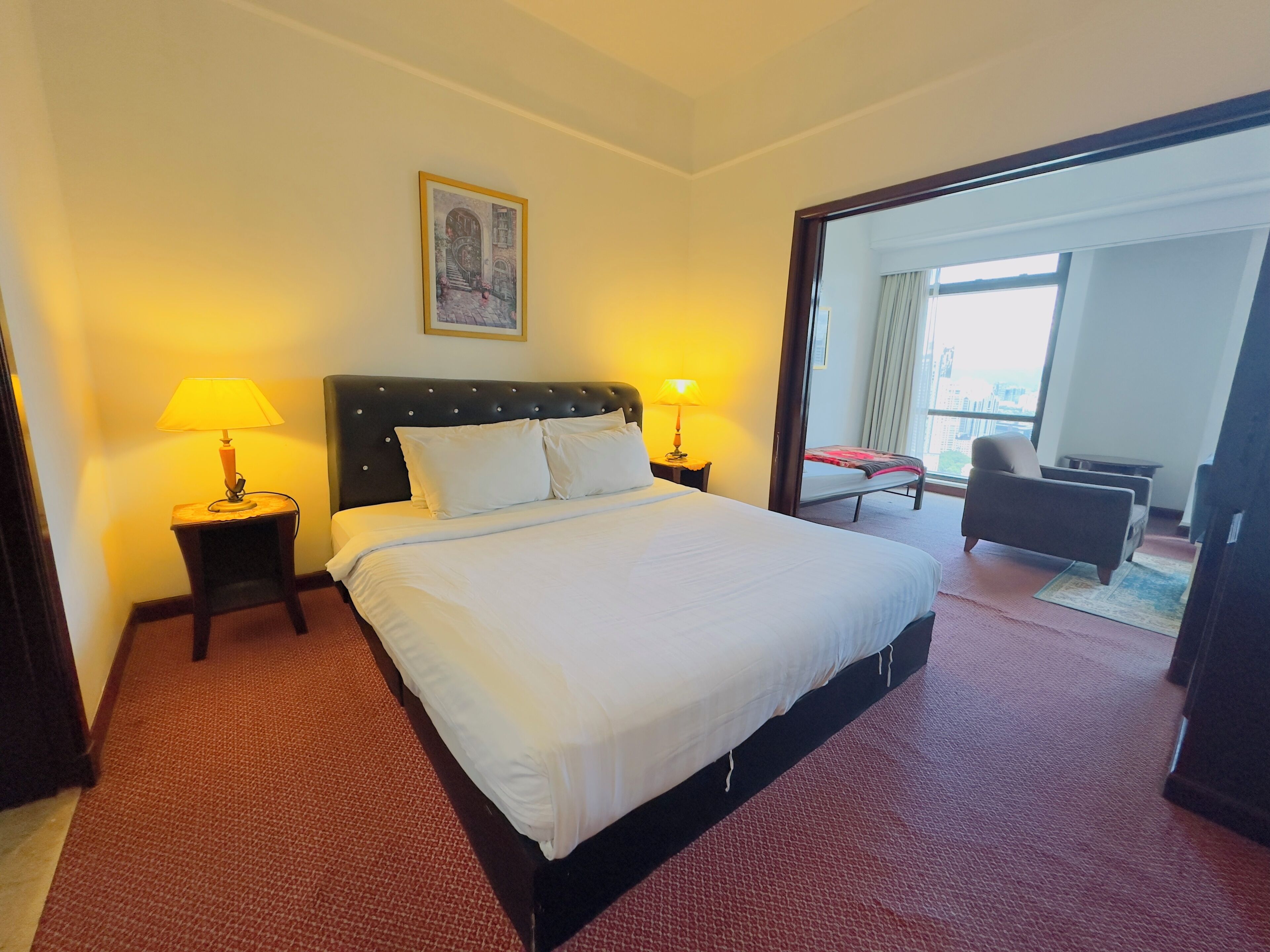 Ein Hotelzimmer mit einem großen Bett, zwei Nachttischlampen, einem gerahmten Bild an der Wand und Blick auf die Stadt durch das Fenster.