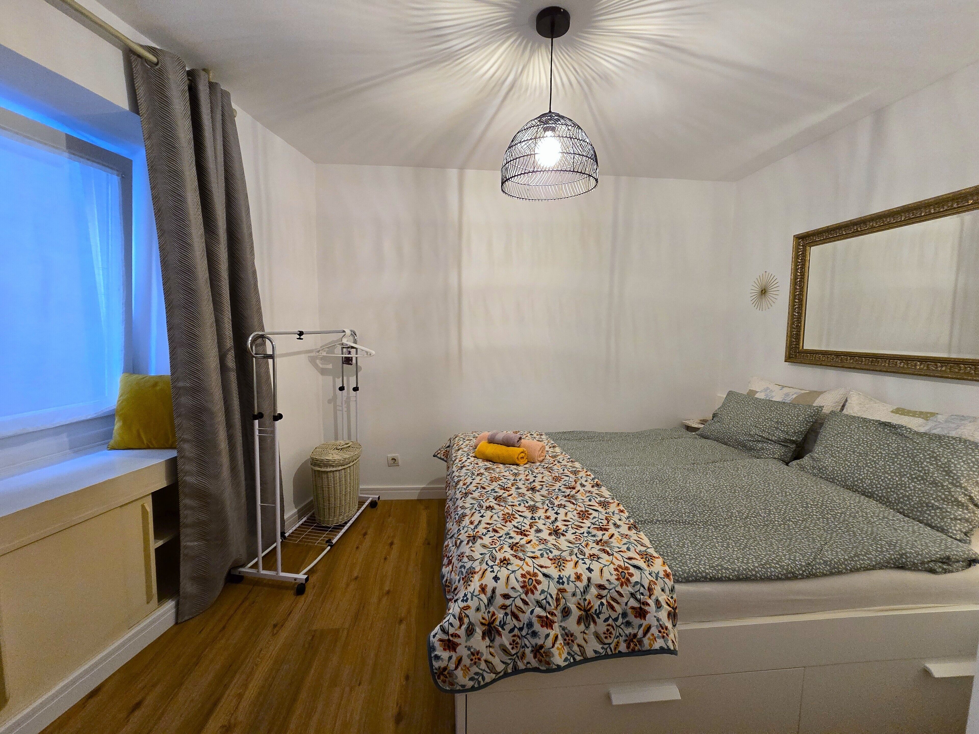A bedroom with a bed, a hanging light fixture, a window with curtains, a bedside table, and a wall-mounted mirror.