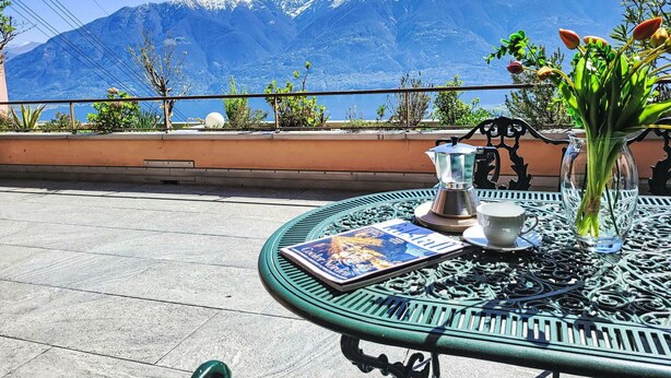 A round metal table with a coffee pot, cup, and a magazine on a balcony overlooking a mountain range. - Beautiful apartment for 4 people with pool, WIFI, TV, patio, pets allowed and panoramic view (Orselina)