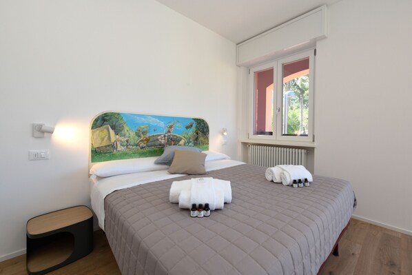 A neatly made bed with a quilted bedspread, a painting above it, and a window with blinds.
