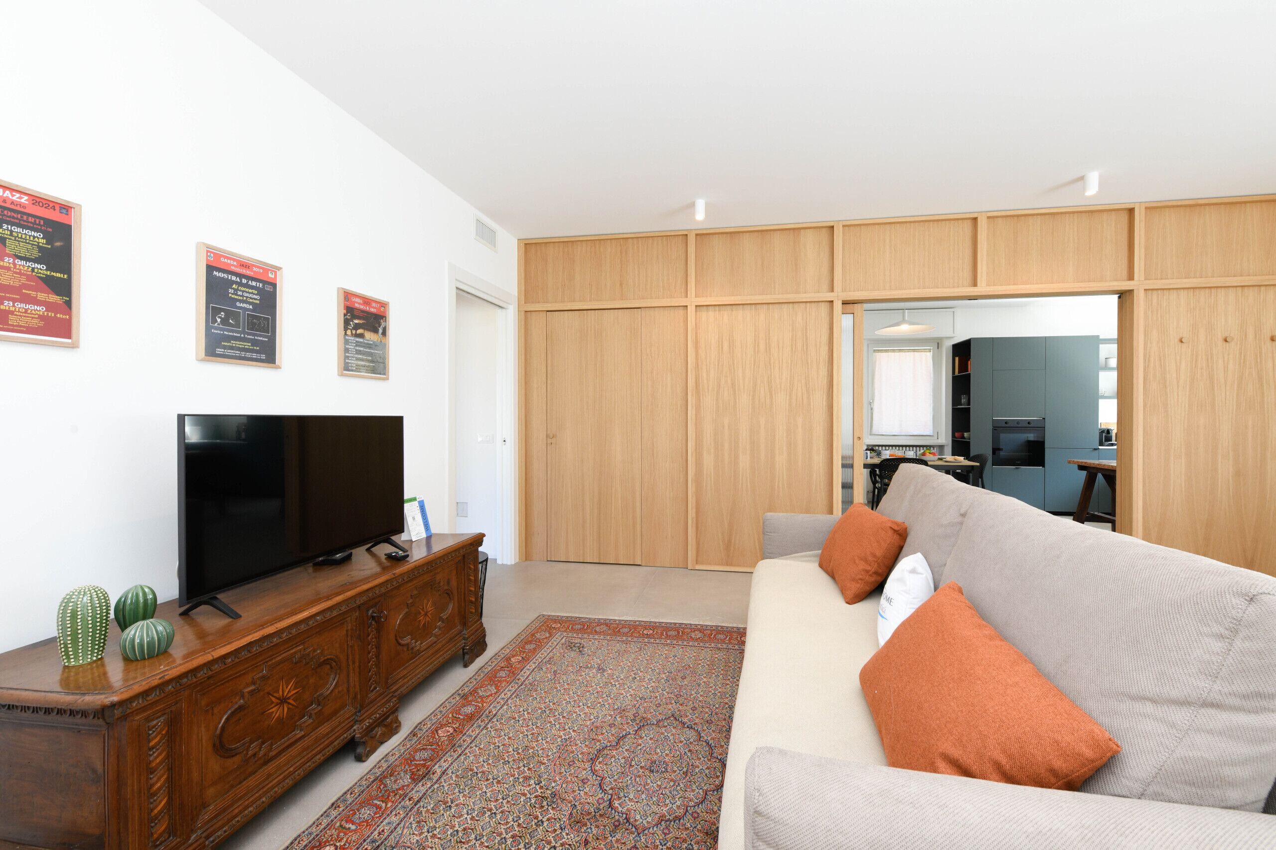 A living room with a flat-screen TV on a wooden cabinet, a grey sofa with orange cushions, and a patterned rug.