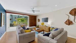 A living room with a sofa, coffee table, and a TV. There's a balcony with plants and a view of greenery.
