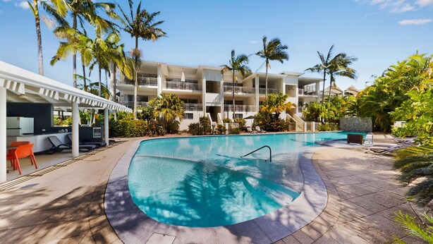A swimming pool with a curved edge, surrounded by palm trees and a multi-story building with balconies.