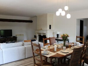 A dining area with a round table set for six, surrounded by wooden chairs. A fireplace is visible in the background, and a television is mounted on the wall to the left.