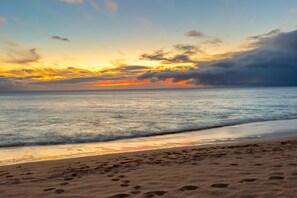 A beach with footprints leading to the water, calm sea, and a sunset sky.