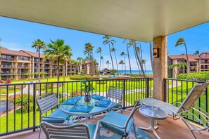 A balcony with a table set for dining, chairs, and a lounge chair, overlooking a pool and palm trees.