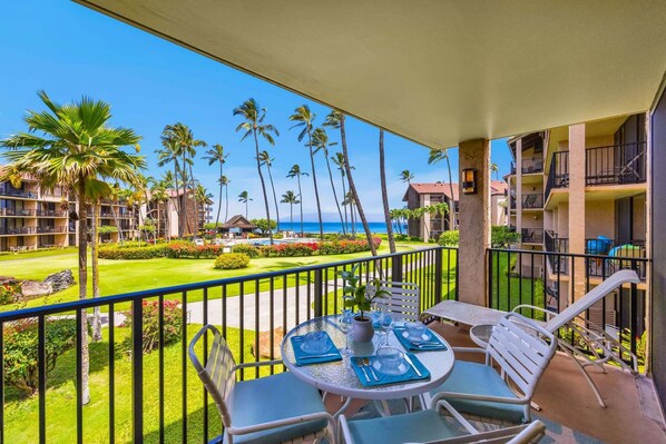 A balcony with a table set for dining, overlooking a well-maintained lawn and palm trees, with a view of the ocean and hotel buildings in the background.