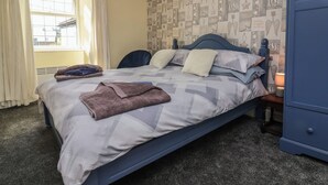 A neatly made bed with a patterned bedspread, pillows, and a folded towel. There is a window with curtains, a radiator, and a blue wardrobe in the room. - Tea Cosy Cottage (Bude)
