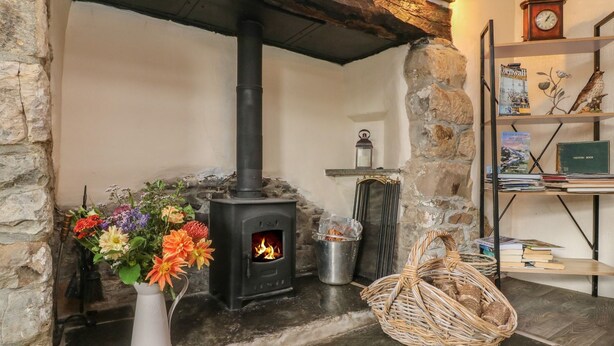 A stone fireplace with a black stove, a white vase with flowers, a metal pot, and a wicker basket on the floor. - Tea Cosy Cottage (Bude)