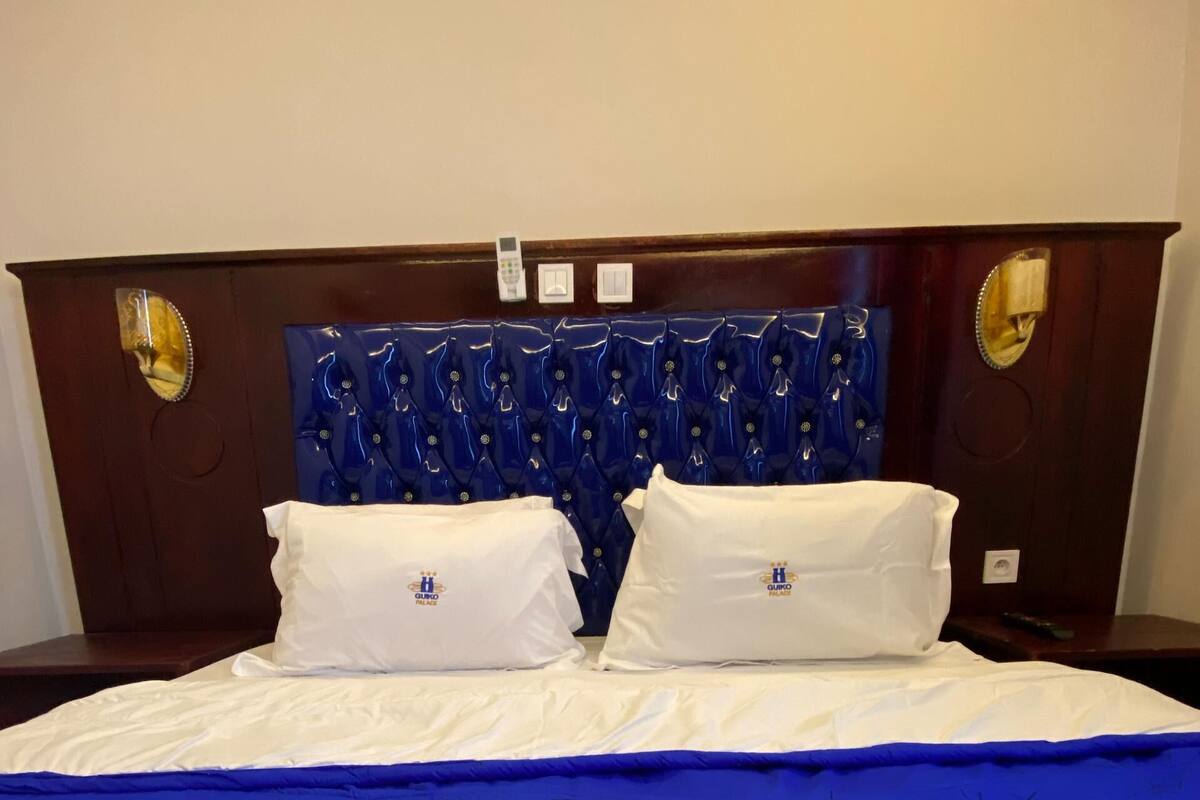 A hotel room with a bed featuring a blue and white bedspread with the hotel logo, a wooden headboard with a blue patterned design, and two white pillows with the hotel logo.