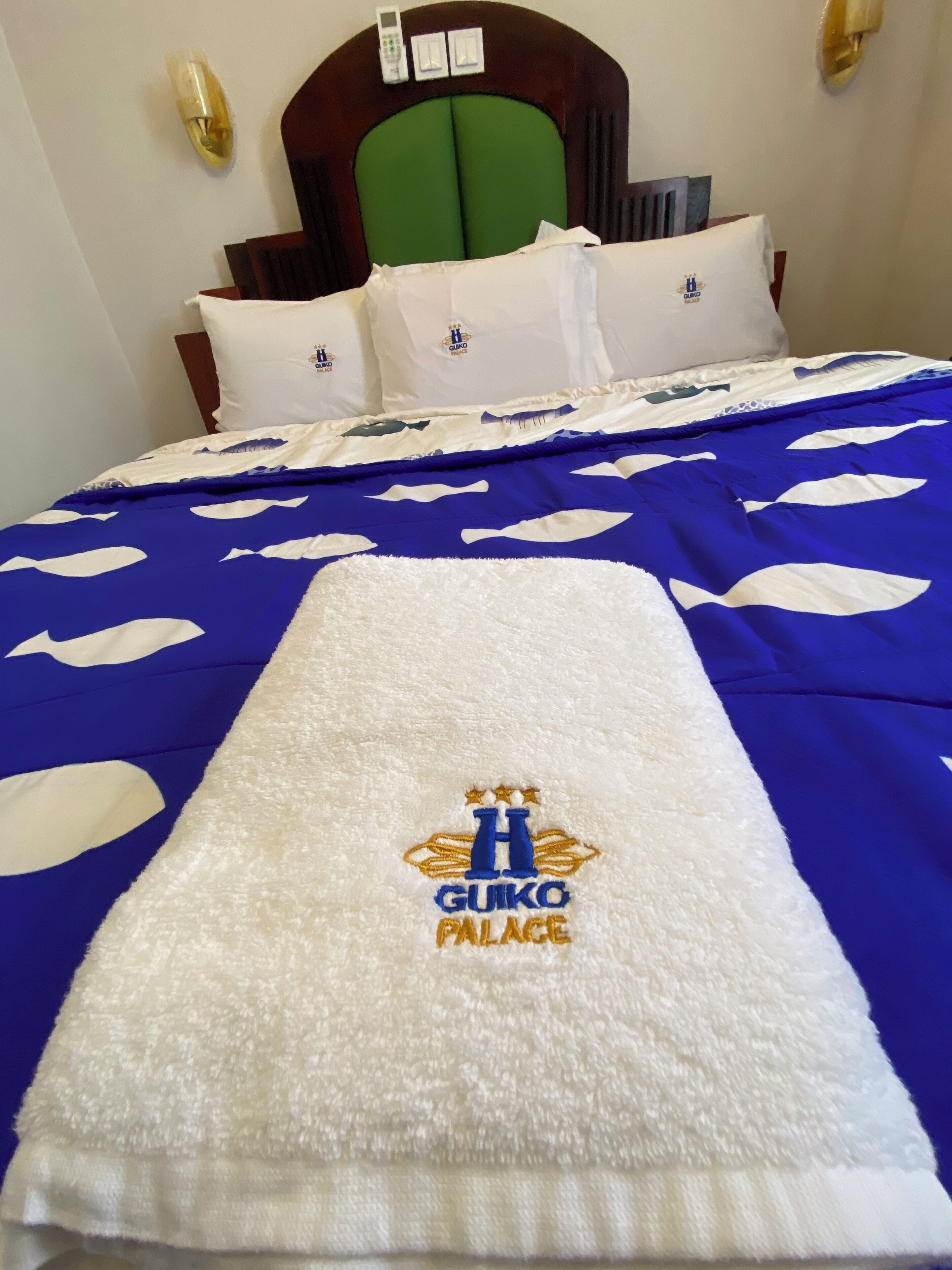 Een hotelkamer met een bed, een groen hoofdeinde, witte kussens en een blauw-witte dekbedovertrek met een handdoek waarop het hotellogo staat.