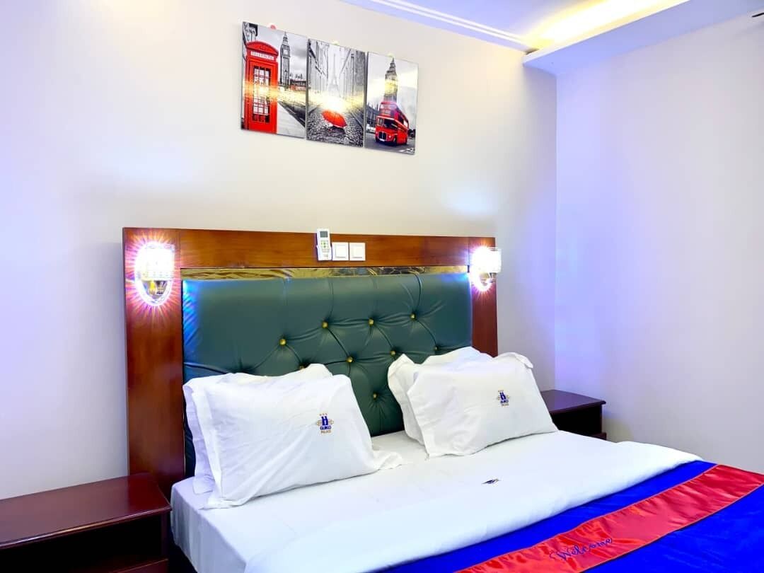 Een hotelkamer met een groen gestoffeerd hoofdeinde, wit beddengoed en een rode en blauwe plaid. Twee witte kussens met een logo, een nachtkastje met een lamp en een aan de muur bevestigd toestel.