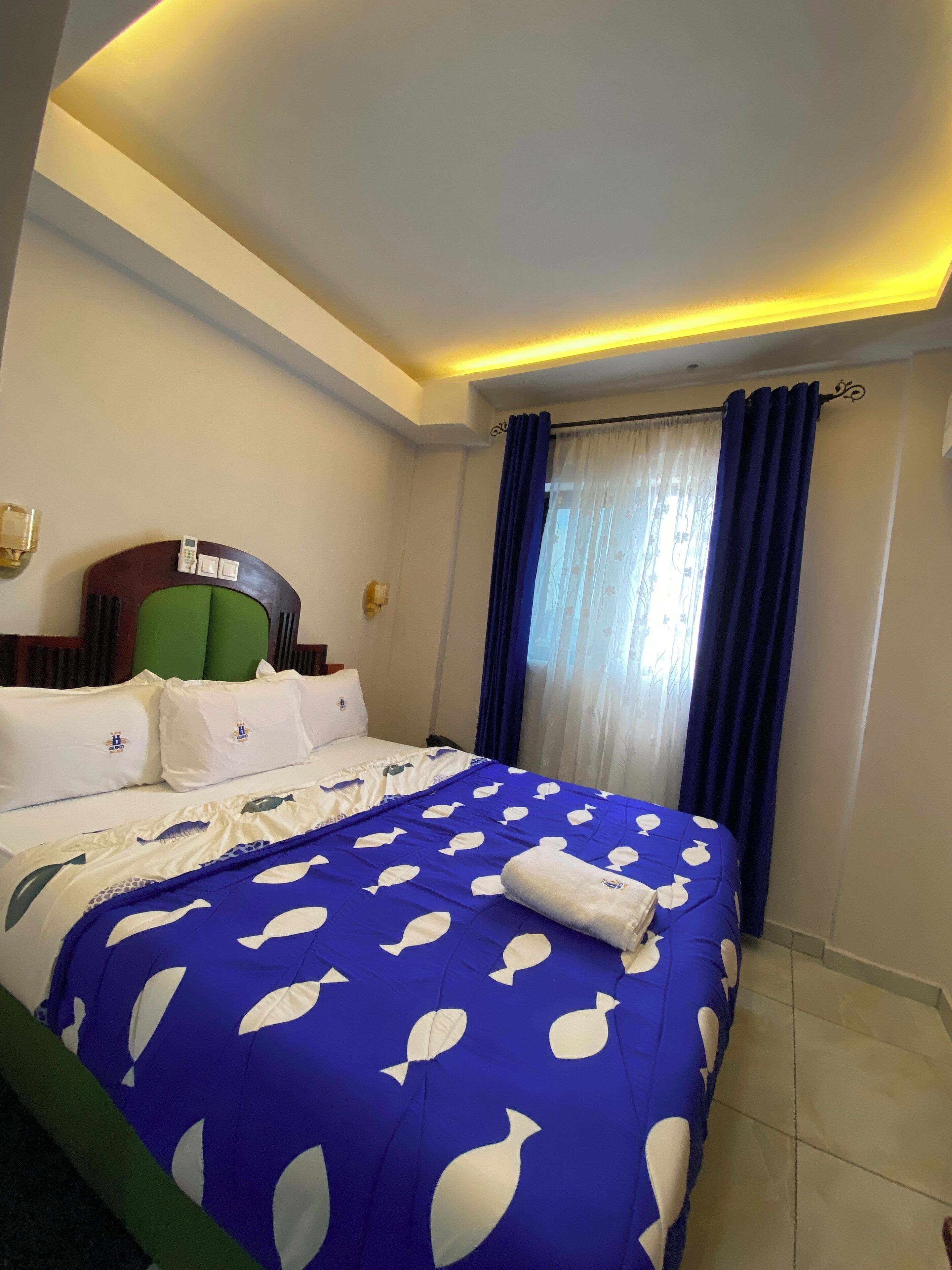 Habitación de hotel con una cama, cabecera verde, teléfono y ventana con cortinas azules.