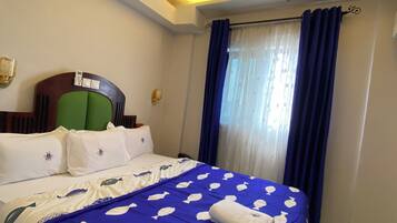 Habitación de hotel con una cama, cabecera verde, teléfono y ventana con cortinas azules.