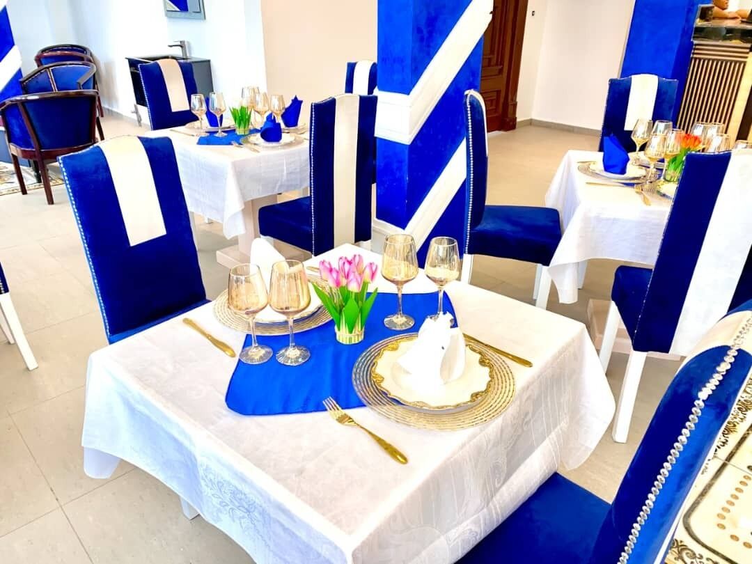 A dining area with tables set for a formal meal, featuring blue and white tablecloths, gold cutlery, and floral centerpieces.