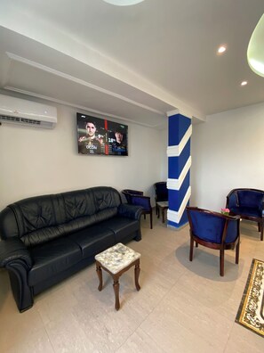 A modern living room with a black leather sofa, two blue armchairs, a small wooden coffee table, and a large flat-screen TV mounted on the wall displaying a sports event. - Guiko Palace Hôtel (Douala)