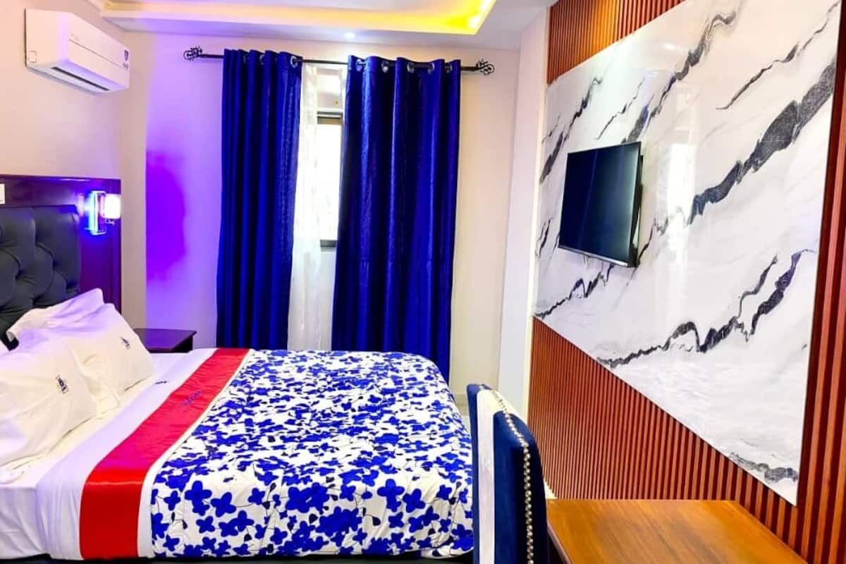 A hotel room with a bed, a TV mounted on the wall, and a window with blue curtains.