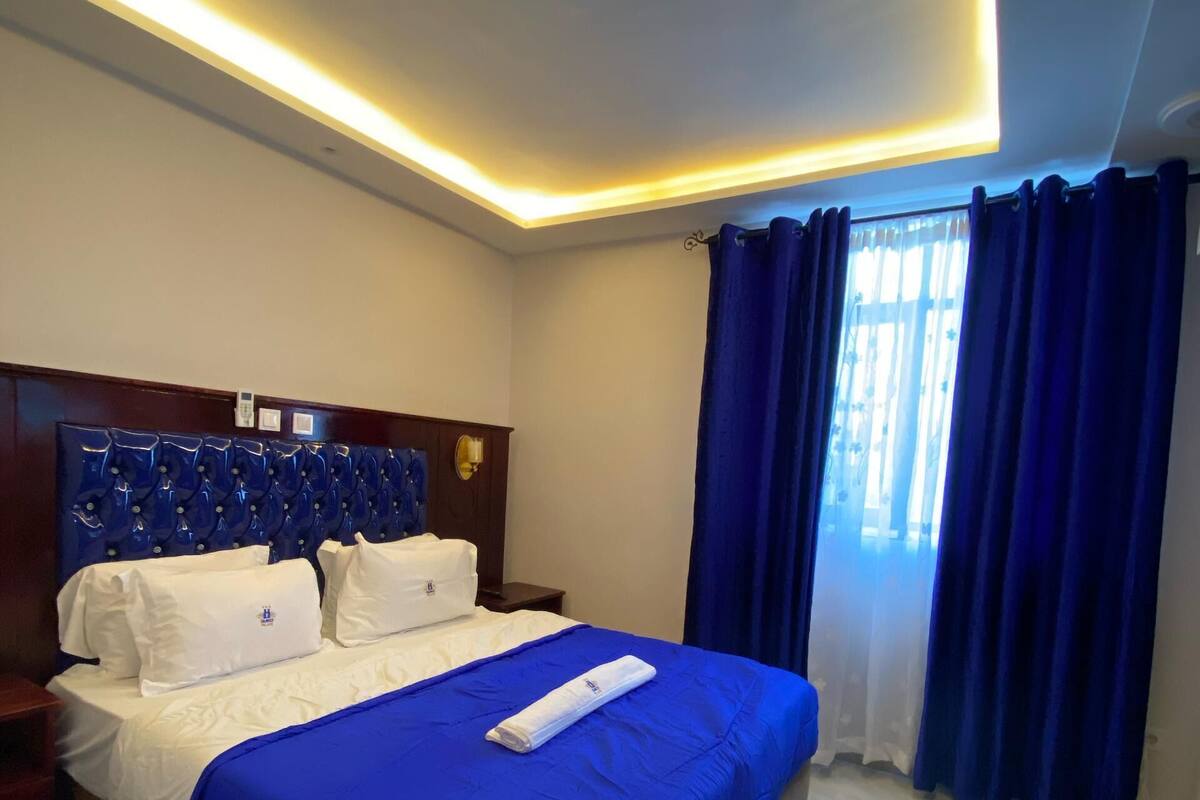 A hotel room with a large bed, a wooden headboard, a bedside table, a nightstand, and a window with blue curtains.