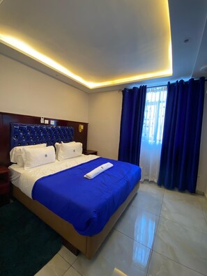 A hotel room with a large bed, a wooden headboard, a bedside table, a nightstand, and a window with blue curtains. - Guiko Palace Hôtel (Douala)
