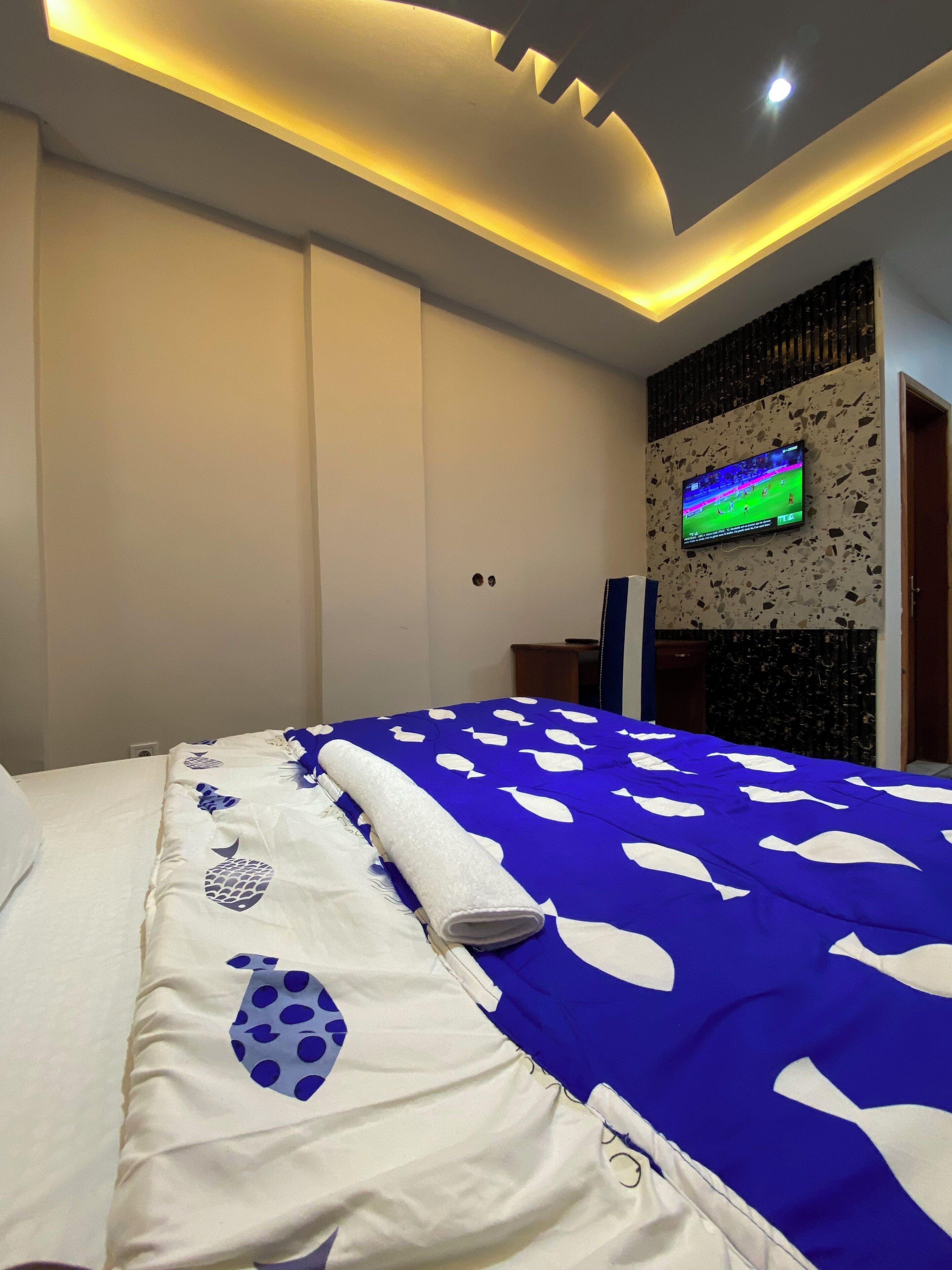 A hotel room with a bed featuring a blue and white bedspread, a flat-screen TV mounted on a textured wall, and a ceiling with recessed lighting.