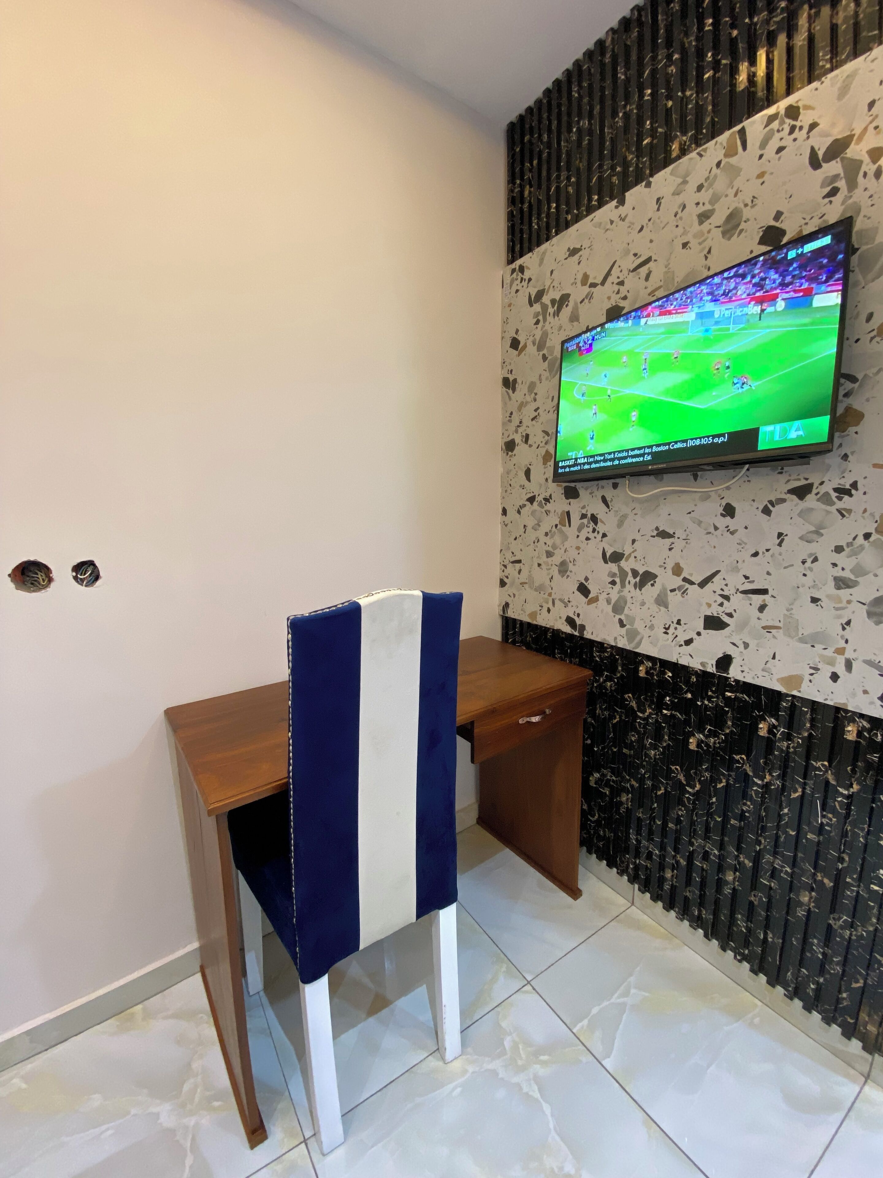 A room with a wooden desk, a chair with a striped cushion, a television mounted on a textured wall, and a tiled floor.