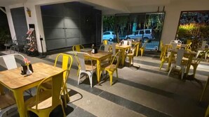 Ein Restaurant mit gelben Stühlen und Tischen, einer großen schwarzen Tür und Blick auf geparkte Autos und Grünflächen vor dem Haus.